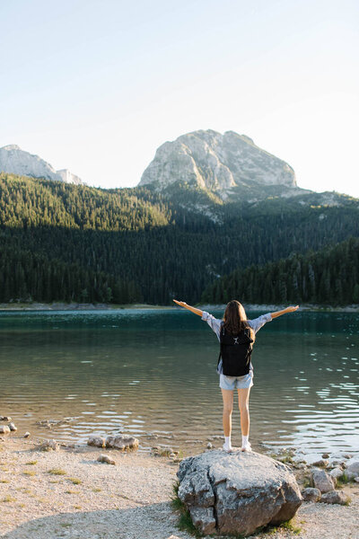 Happy woman tourist stands on a large stone with raised hands. The tourist has a black hiking backpack on his back. Against the backdrop of mountains, forest and mountain lake. Back view. Hiking.