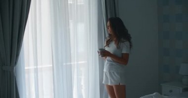 Yatağındaki güzel kadın dinlendi ve pencerelerdeki perdeleri açtı ve sabah yatak odasında güneş ışığı altında temiz hava almak için bir fincan kahveyle pencereden dışarı baktı..