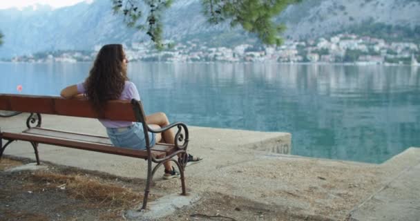 Glad kvinna turist med lockigt hår ser på bänken och har den vackra utsikten över den blå bukten och bergen. Tidig morgon. Bakifrån — Stockvideo