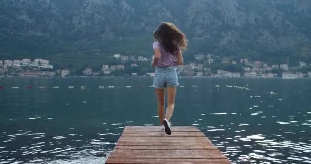 Jovem turista feliz com cabelo encaracolado corre ao longo do cais de madeira, pára e aperta as mãos. Um sentido de liberdade — Vídeo de Stock