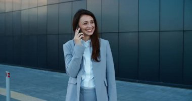 Takım elbiseli gülümseyen iş kadını modern bir binanın yanında duruyor ve cep telefonuyla konuşuyor. Kadın portresi
