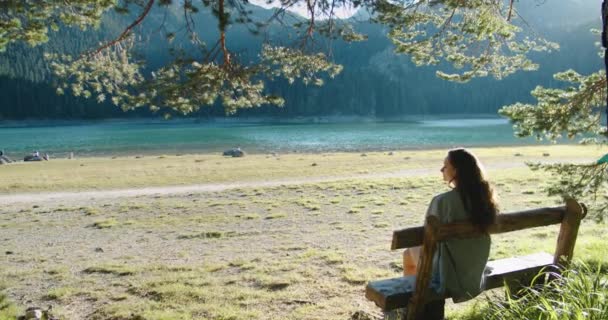 Mujer feliz turista se sienta en un banco y disfruta de una hermosa vista de un lago de montaña, bosque y montañas. Puesta de sol. Vista trasera — Vídeos de Stock