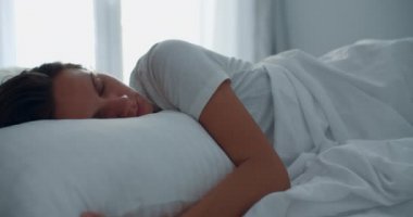 Genç bir esmer kadın, sabah yatakta büyük bir pencerenin arka planında uyanır. Gülümseyen bir kadının portresi