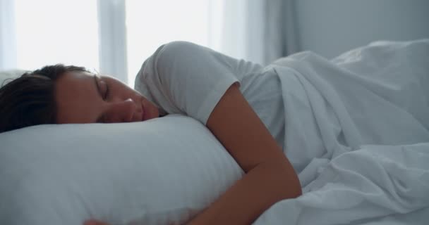 Eine junge brünette Frau wacht morgens vor dem Hintergrund eines großen Fensters im Bett auf. Porträt einer lächelnden Frau — Stockvideo