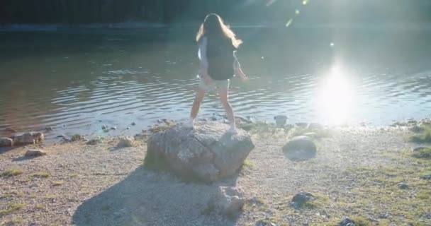 Donna felice turista con un grande zaino salta su una grande pietra che si affaccia sul lago e sulle montagne, si erge sulla pietra e guarda la natura. Vista posteriore — Video Stock