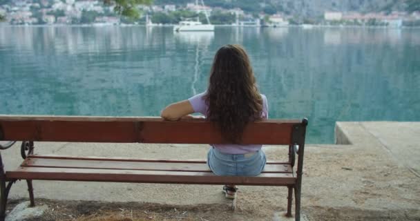 Turista mulher feliz com cabelo encaracolado senta-se em um banco e examina a natureza. Vista mar e montanha. Visão traseira — Vídeo de Stock
