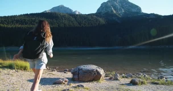 큰 가방을 들고 호수와 산이 내려다 보이는 큰 돌 위에 뛰어 올라서 자연을 바라보는 행복 한 여자가 있다. 뒷모습 — 비디오
