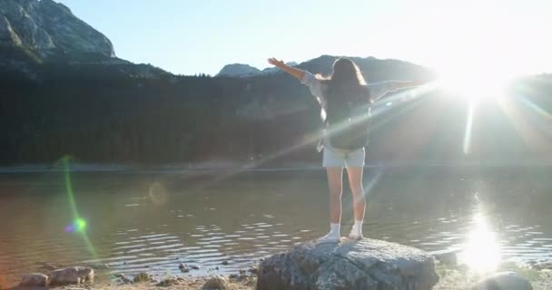 Mujer feliz turista con una gran mochila salta sobre una gran piedra con vistas al lago y las montañas, se encuentra en la piedra y mira a la naturaleza. Vista trasera — Vídeos de Stock