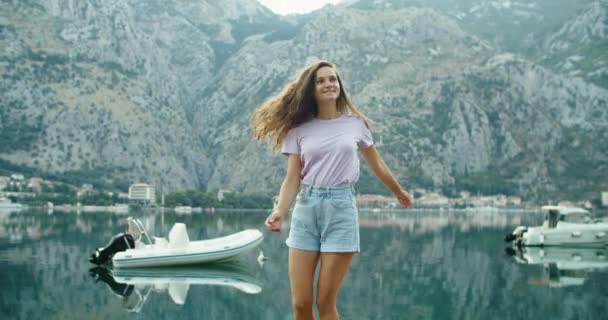 Szczęśliwa młoda kobieta turysta biegnie po drewnianym molo i patrzy na piękny widok. Zatoka i góry. Łodzie i nożyce stoją na wodzie — Wideo stockowe