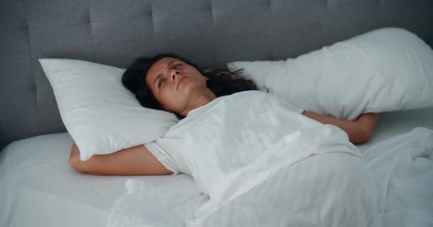 Раздраженная женщина проснулась рано утром от постороннего шума. Женщина покрывает себя подушками, чтобы не слышать шума в спальне рано утром — стоковое видео