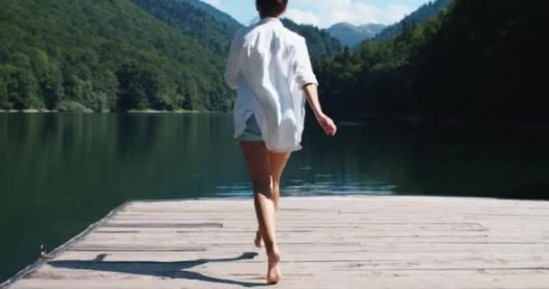 하얀 셔츠를 입고 기뻐하는 한 여자는 나무로 된 교각을 맨발로 걸어 올라가 자연의 아름다운 경관을 즐긴다. 산과 호수의 전경. 뒷모습 — 비디오