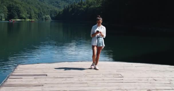 Happy brunette woman walks on a wooden pier near a mountain lake and prints messages on a mobile phone. Female portrait. Mountain and lake views. — Stock Video