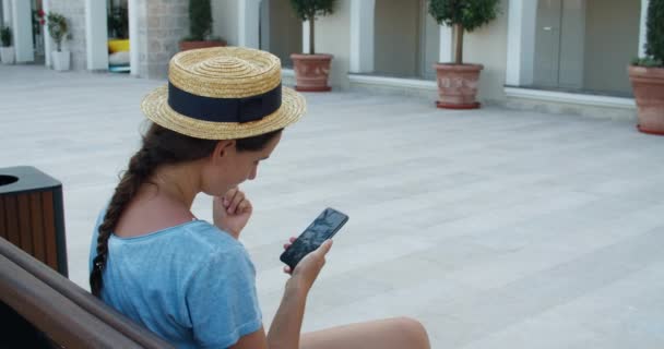 Une femme heureuse portant un chapeau est assise sur un banc de la ville et lit un message sur un téléphone portable. Vue latérale — Video