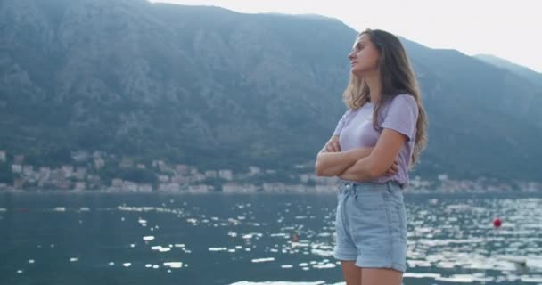 Šťastná brunetka žena stojí na molu, usmívá se a má výhled na přírodu. Pohled na záliv a hory. — Stock video
