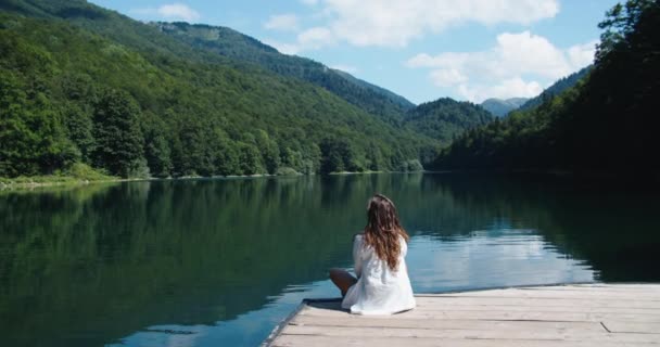 행복 한 여자는 나무로 된 부두에 앉아 아름다운 자연을 즐긴다. 호수와 산의 경치가 좋다. 뒷모습 — 비디오