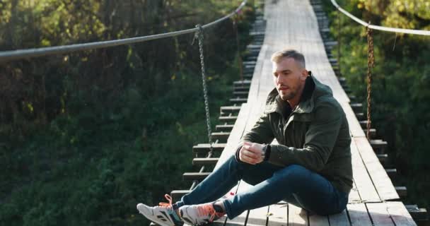 Un jeune touriste en vêtements chauds s'assoit sur un pont suspendu en bois et regarde la nature, se réchauffe les mains. Plantes vertes — Video