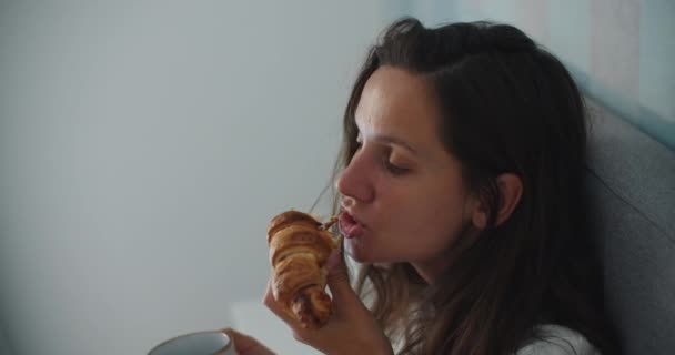 Uma mulher morena está tomando café da manhã com um croissant e café no início da manhã. Close-up retrato de uma mulher — Vídeo de Stock