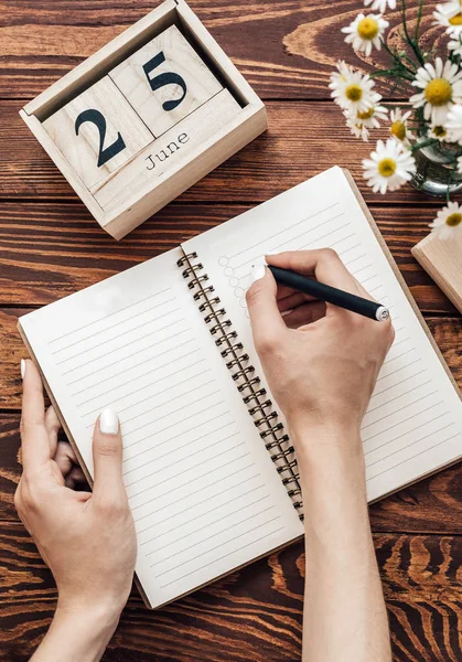 A young woman makes a shopping list or a list of plans in a notebook on a wooden table. conceptual photo