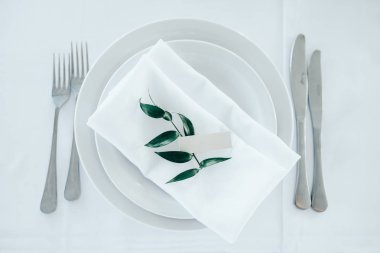 Beyaz masa örtüsü üzerinde çatal ve bıçak olan beyaz bir tabak. Plakadaki yeşil bitki ve isim plakası