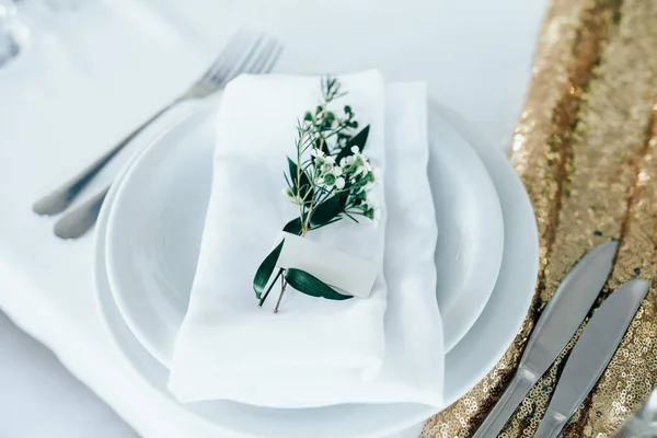 Beyaz masa örtüsü üzerinde çatal ve bıçak olan beyaz bir tabak. Plakadaki yeşil bitki ve isim plakası