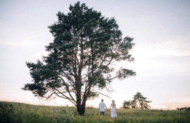 Sevgi dolu çift gün batımının arka planında büyük, güzel bir ağaçta el ele yürüyorlar. Kavramsal güzel aşk fotoğrafı. Yazıtlar için yer var.