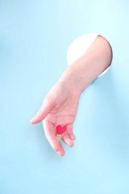 Close up of human hand protruding through hole in blue background, holding red heart. clipart