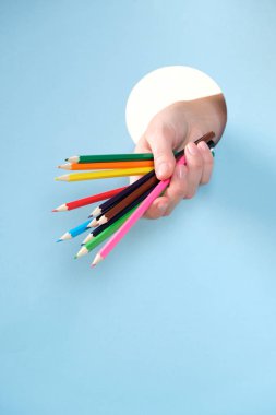 Human hand protruding through hole in blue background, holding colored pencil. clipart