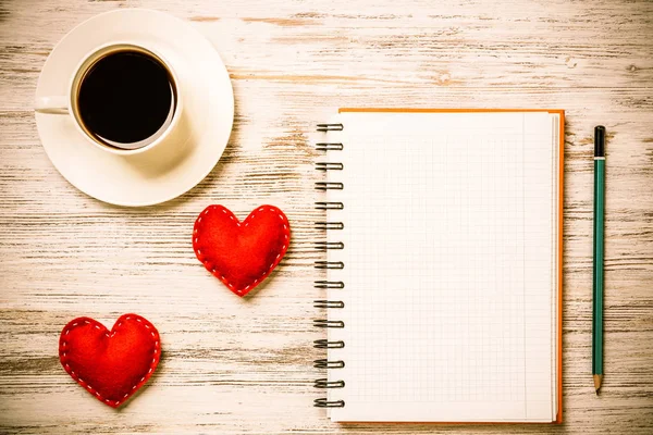 Coffee cup notepad pencil and two red hearts on wooden surface