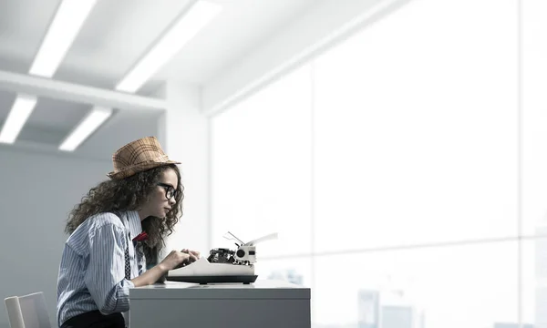Young woman writer in hat and eyeglasses using typing machine while sitting at the table indoors with office view on background.