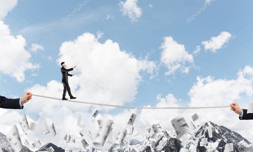 Businessman walking blindfolded on rope among flying documents and above high mountains as symbol of hidden threats and support. Nature view on background.