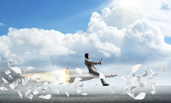 Conceptual image of young businessman in suit flying on rocket above asphalt road and among flying papers with blue sky on background.