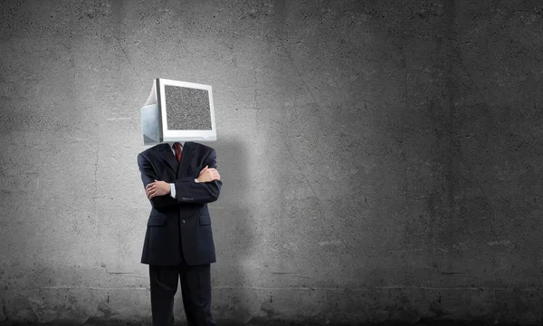 stock image Businessman in suit with monitor instead of head keeping arms crossed while standing inside empty room with gray dark wall on background.