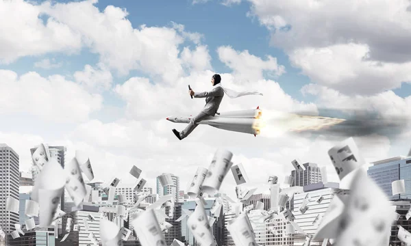 Conceptual image of young and happy businessman in suit flying on rocket among flying papers with modern cityscape with skyscrapers and blue sky on background.