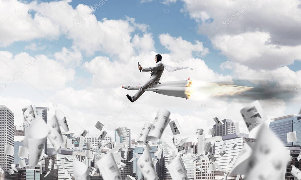 Conceptual image of young and happy businessman in suit flying on rocket among flying papers with modern cityscape with skyscrapers and blue sky on background.