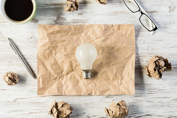 Top view of modern creative workplace with lightbulb on creased piece of sheet, crumpled paper balls and cup of coffee placed on wooden table. Concept of finding of outstanding idea.