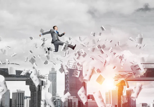 Businessman jumping over gap with flying paper documents in concrete bridge as symbol of overcoming challenges. Sunlight and cityscape on background. 3D rendering.