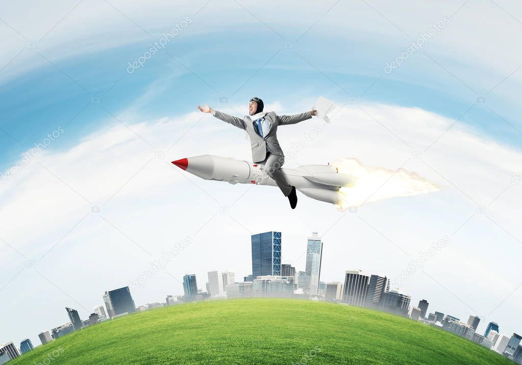 Conceptual image of young businessman in suit flying on rocket with cityscape and blue sky on background.