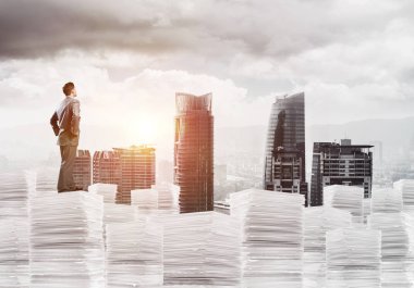 Takım elbiseli kendine güvenen genç işadamı şehir manzarası ve arka planda güneş ışığı olan bir yığın belgenin üzerinde duruyor. Karışık ortam.