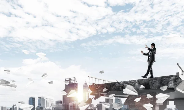 歩くビジネスマンは隠された脅威とリスクのシンボルとして巨大なギャップを持つコンクリート橋に紙飛行機を飛行中で目隠し 都市の景観と背景に日光 レンダリング — ストック写真