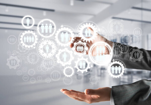 Business woman in black suit keeping white social gear icons in hands with office view on background. Mixed media.