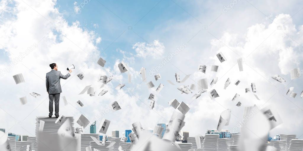 Businessman in suit standing among flying papers with speaker in hand and with skyscape on background. Mixed media.