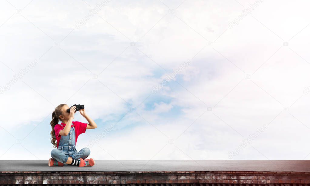 Cute girl of school age on building roof looking in binoculars