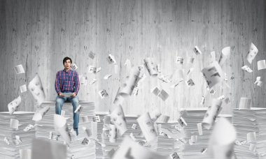 Kağıt belgeleri ile gri duvar arka plan üzerinde uçan arasında casual giyim oturan genç adam. Karışık teknik.