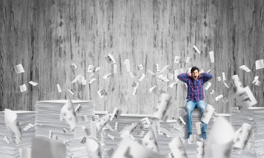 Kağıt belgeleri ile gri duvar arka plan üzerinde uçan arasında casual giyim oturan genç adam. Karışık teknik.