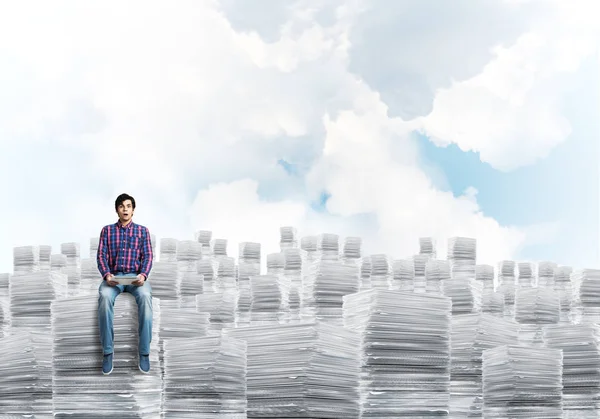 カジュアルウェアの背景に Cloudly 天空と書類の山に座っている若い男 ミクスト メディア — ストック写真