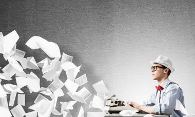 Genç yazar şapka ve gözlük arka plan üzerinde uçan kağıtları arasında ve gri beton duvara karşı masada otururken yazarak makinasıyla adam.