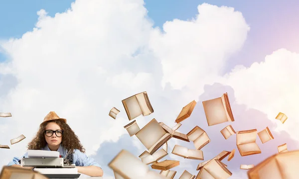 Young and beautiful woman writer in hat and eyeglasses using typing machine while sitting at the table among flying books with cloudy skyscape on background.