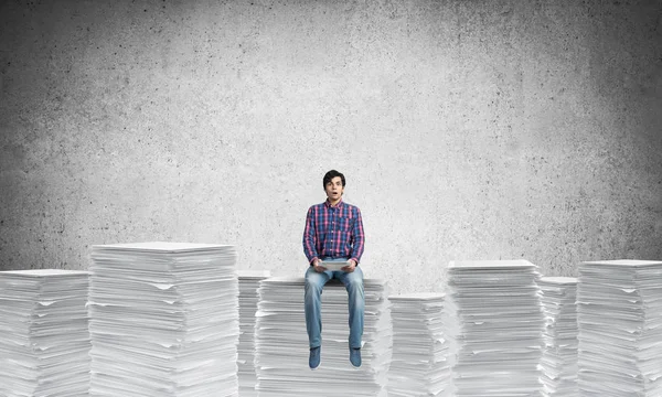 カジュアルな服の背景に灰色の壁と書類の山に座っている若い男 ミクスト メディア — ストック写真
