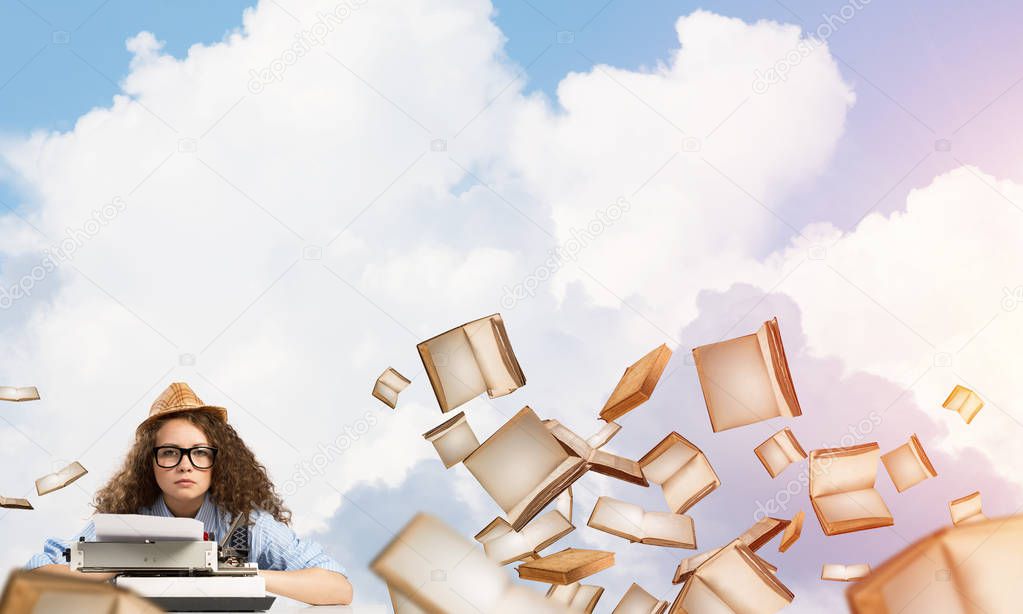 Young and beautiful woman writer in hat and eyeglasses using typing machine while sitting at the table among flying books with cloudy skyscape on background.