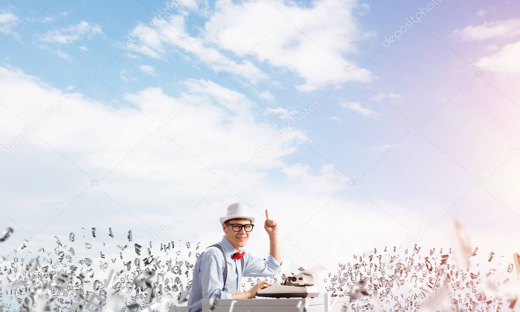 Young man writer in hat and eyeglasses using typing machine and pointing upside while sitting at the table among flying letters with cloudy skyscape on background.